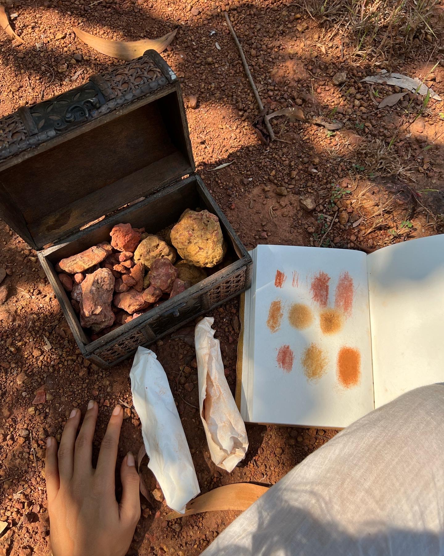 A wooden box of ochre and red pigment rocks beside a sketchbook of earth swatches