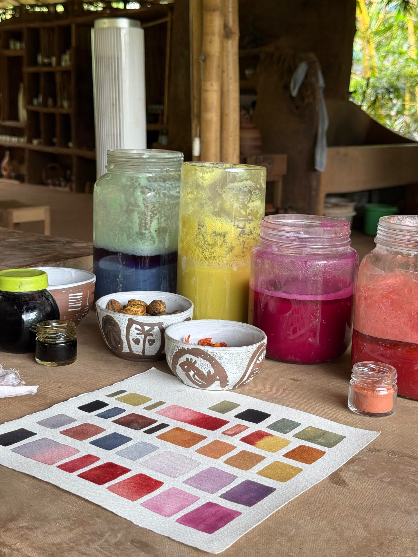 Jars of plant dyes in green, yellow, pink and red beside a painted colour chart